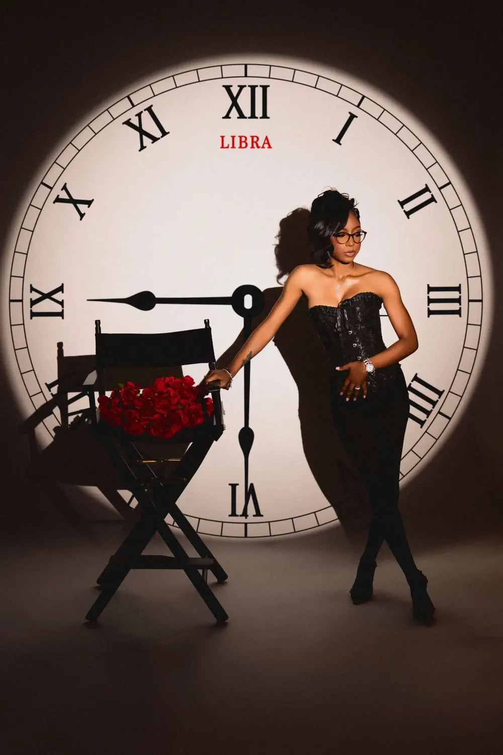 Woman in a black corset leaning on a director's chair with roses in front of a projected clock face