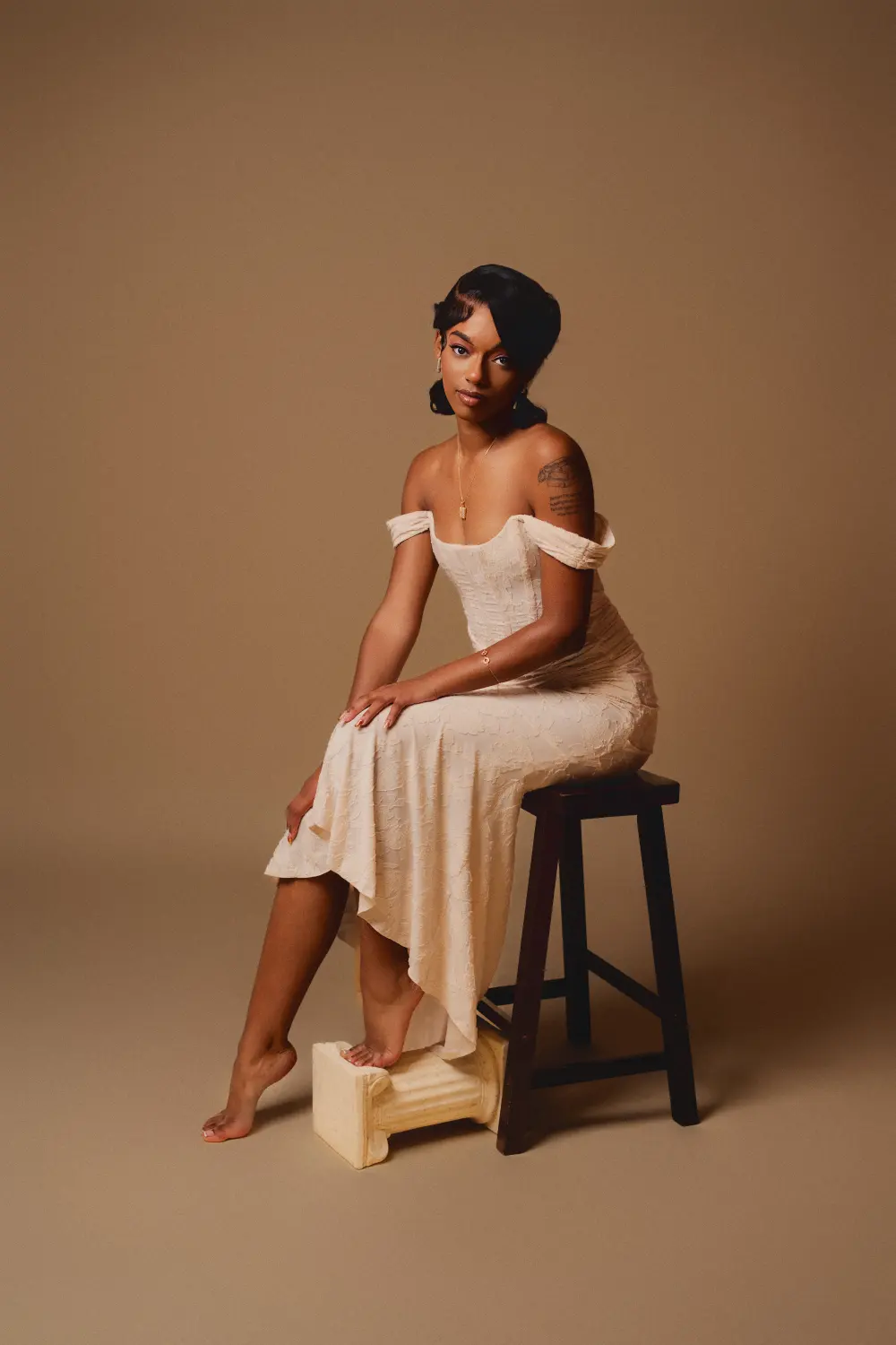 Woman in an off-shoulder cream dress seated on a stool against a tan backdrop