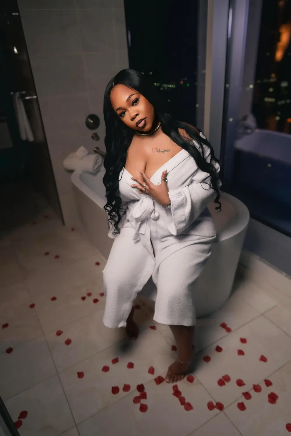 Woman in a white robe sitting on the edge of a hotel bathtub with rose petals on the floor