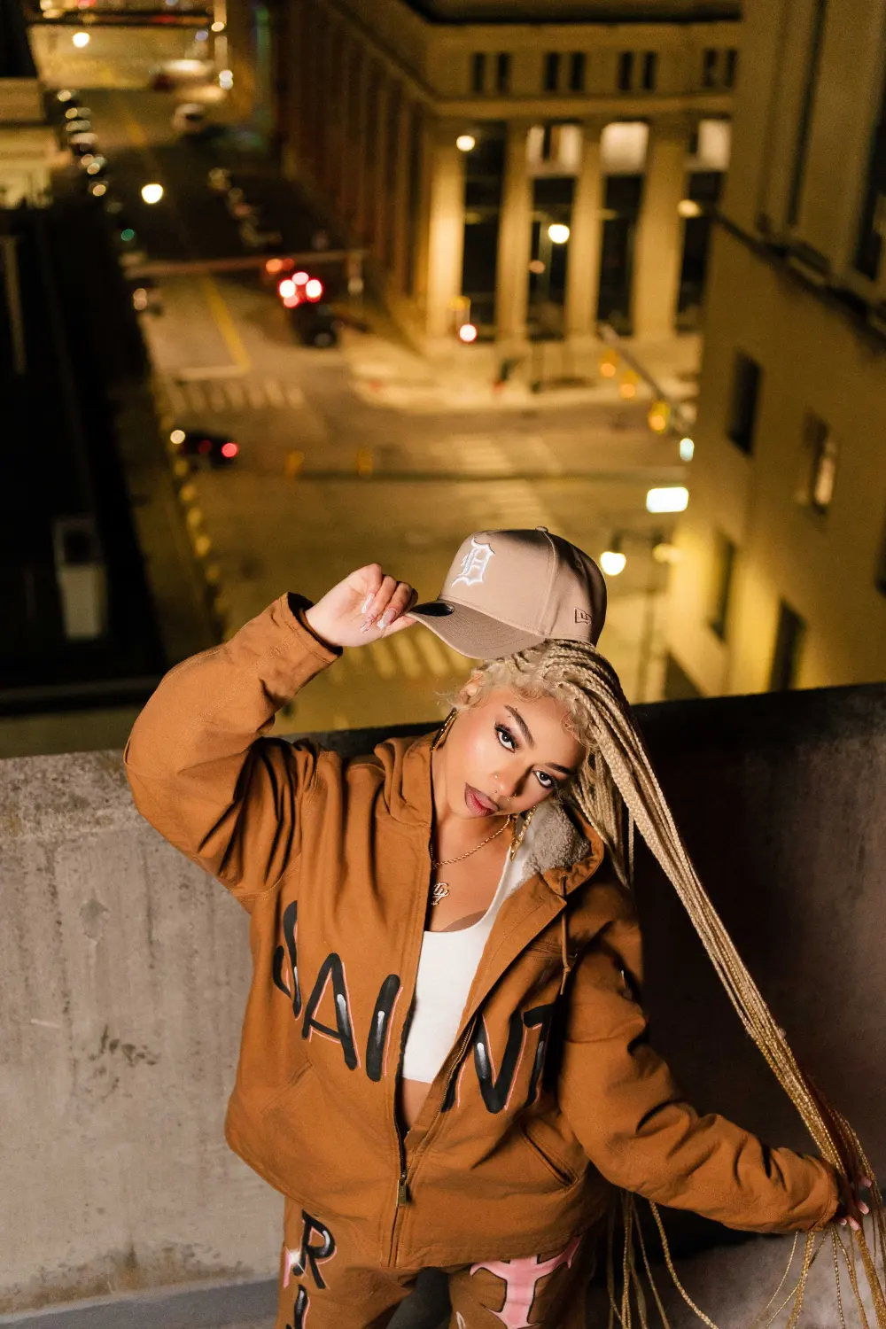 Woman in a brown jacket and cap posing on a parking garage roof above a downtown street at night