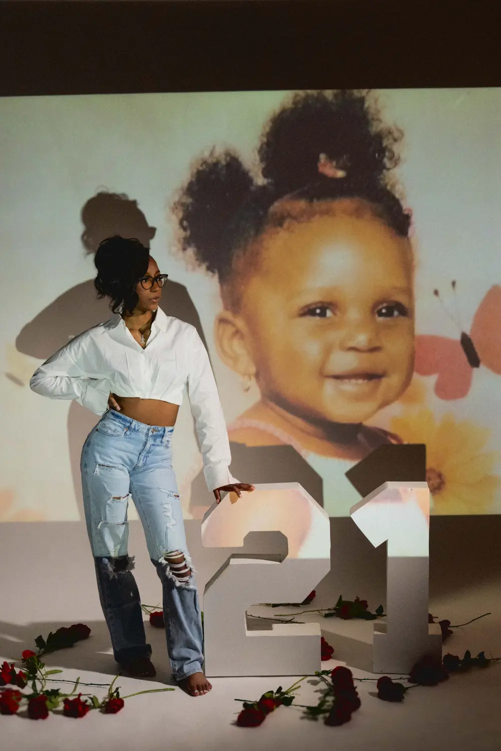 Woman in a white shirt and jeans beside large white 21 numbers and scattered roses, her baby photo projected on the wall behind her