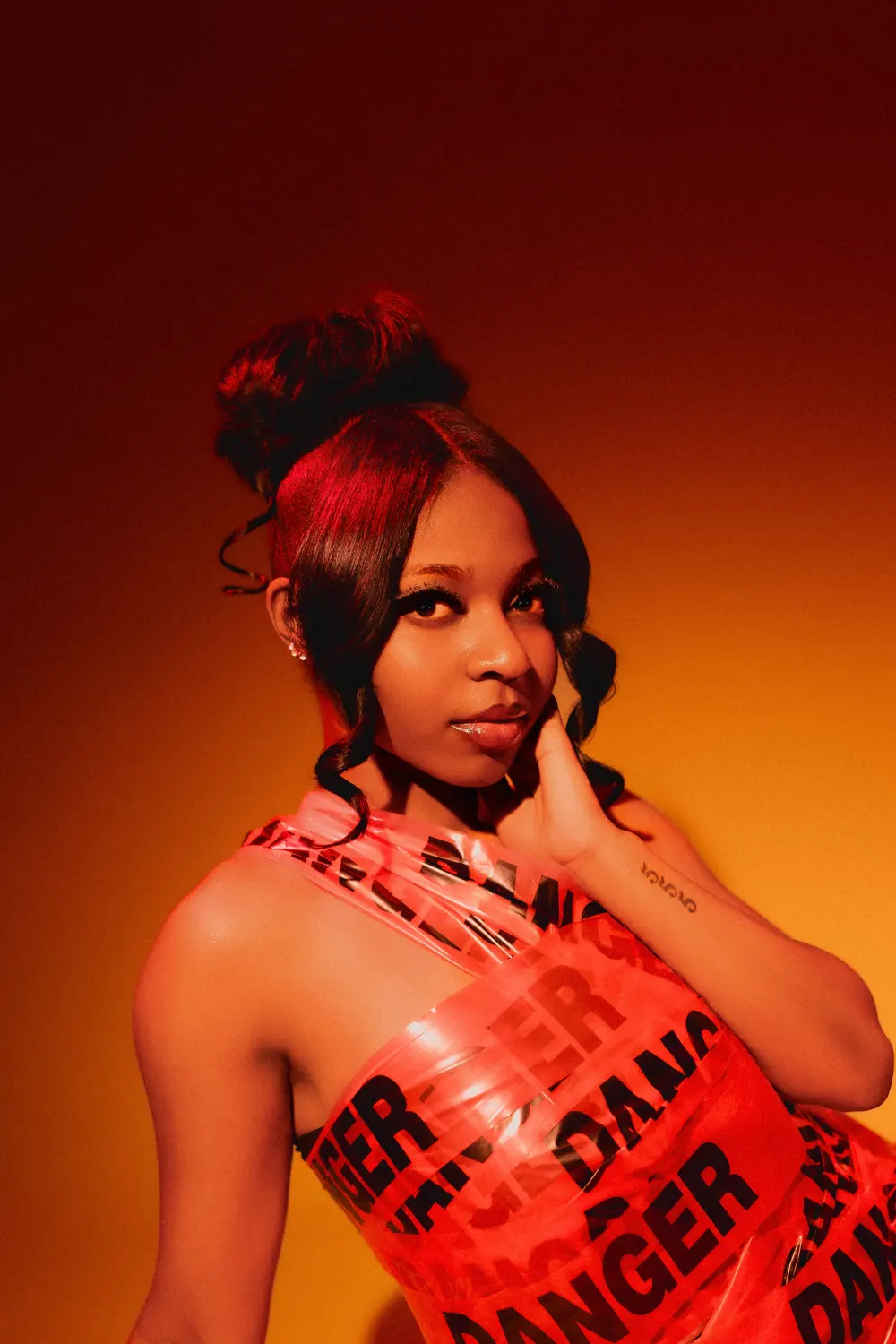 Close portrait in a danger tape dress, hand at her jaw, in warm red light