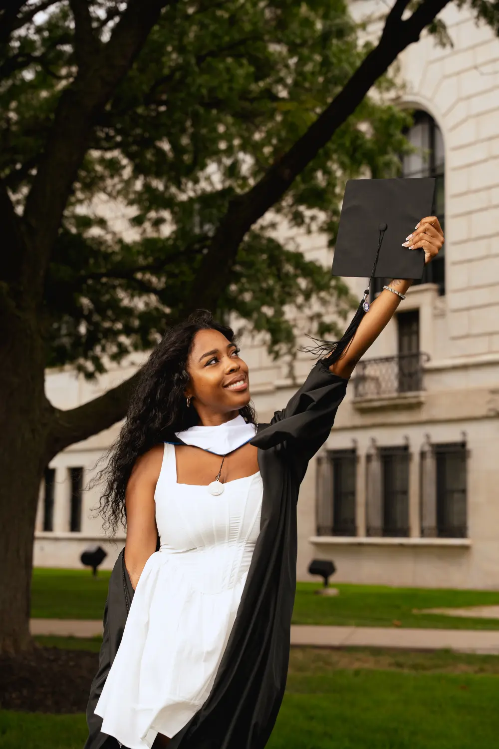 Graduate in a white dress and black gown holding her cap up under the trees outside a stone building