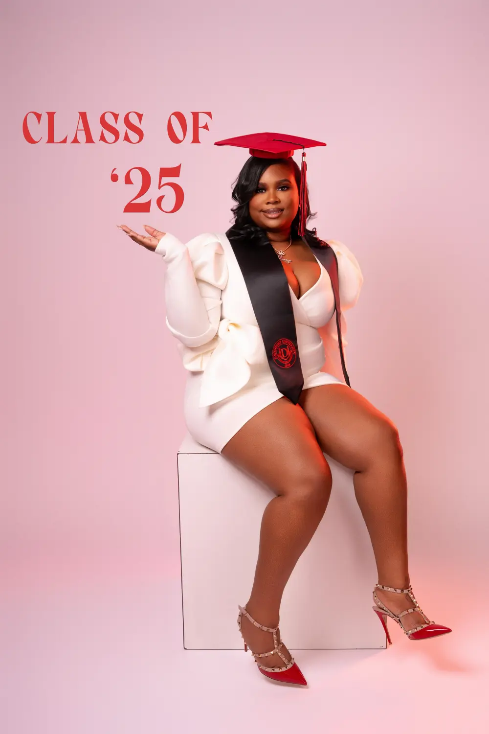 Graduate in a red cap, white dress and black stole seated on a white cube, CLASS OF '25 lettered on the pink wall behind her