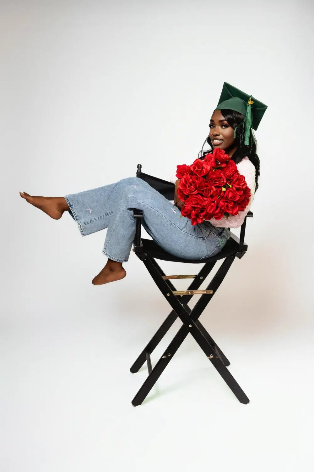 Graduate in a green cap, pink sweater and jeans leaning back in a director's chair with a bouquet of red roses
