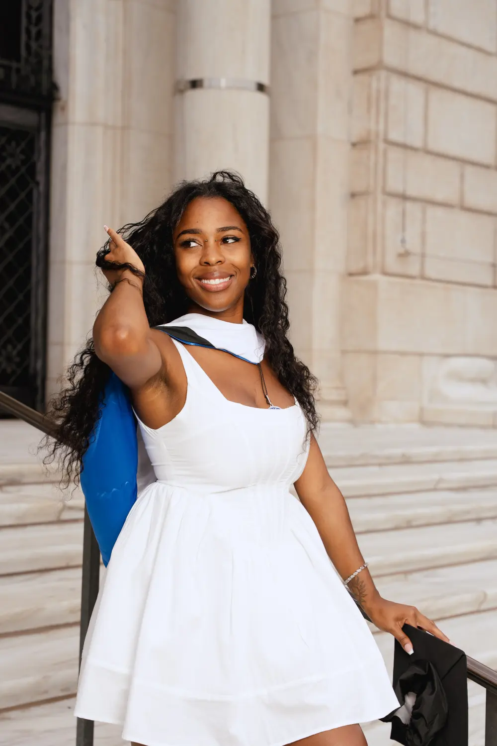 Graduate in a white dress with a blue-lined hood smiling on marble steps downtown, one hand in her hair
