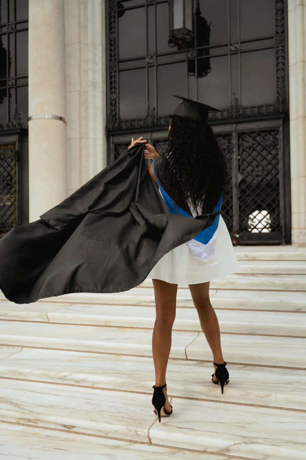 Graduate on white marble steps swinging her black gown out behind her, seen from the back