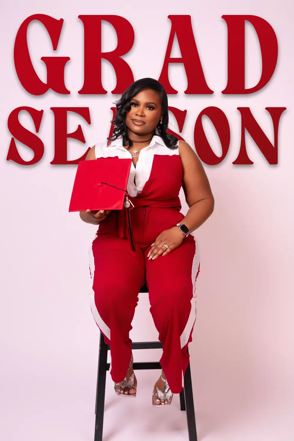 Graduate in a red and white jumpsuit on a stool holding a red cap, GRAD SEASON in big red letters behind her