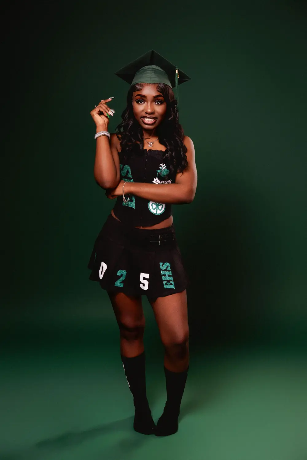 Graduate in a green cap and a black outfit with green lettering, one hand at her face, against a green backdrop