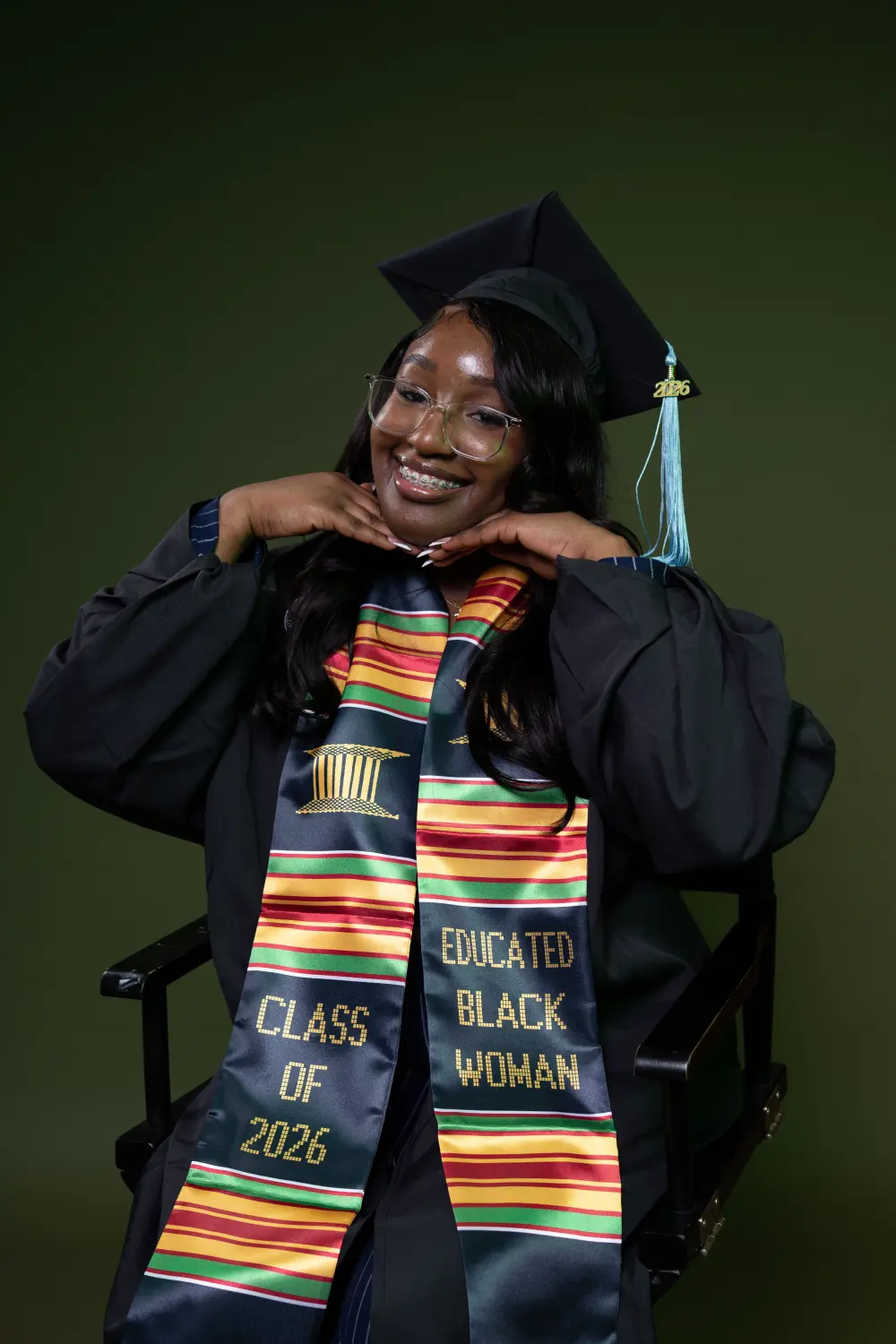 Graduate in glasses and a kente stole smiling with her chin on her hands against a dark green backdrop
