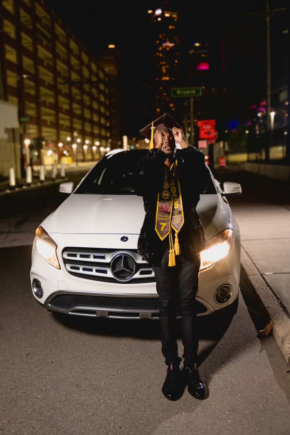 Graduate in a cap and gold stole standing in front of a white car on a downtown street at night