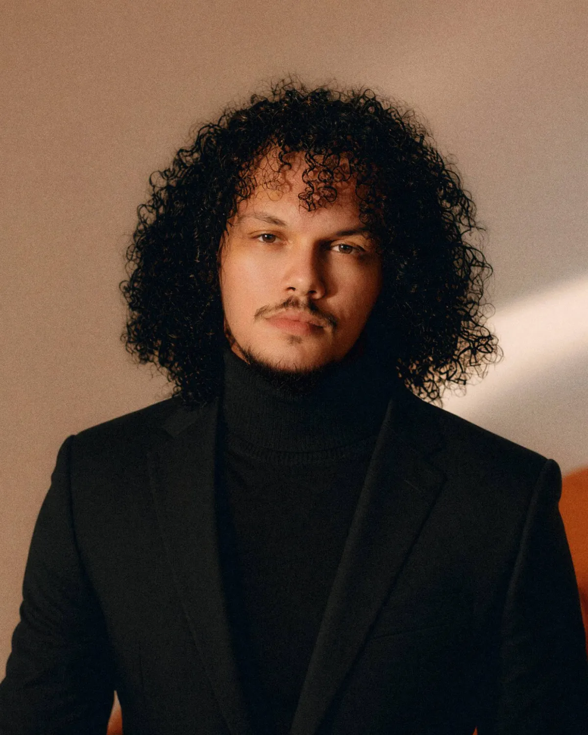 Joey in a black turtleneck and blazer against a warm wall, an orange chair behind him