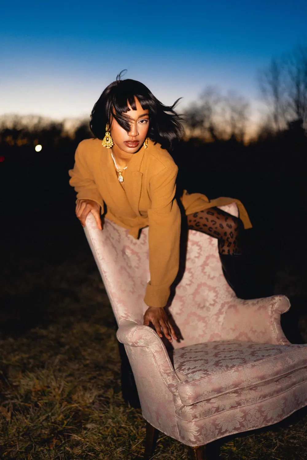 Woman in a camel coat leaning over the back of a pink armchair, her hair flying, a sunset glow behind the trees