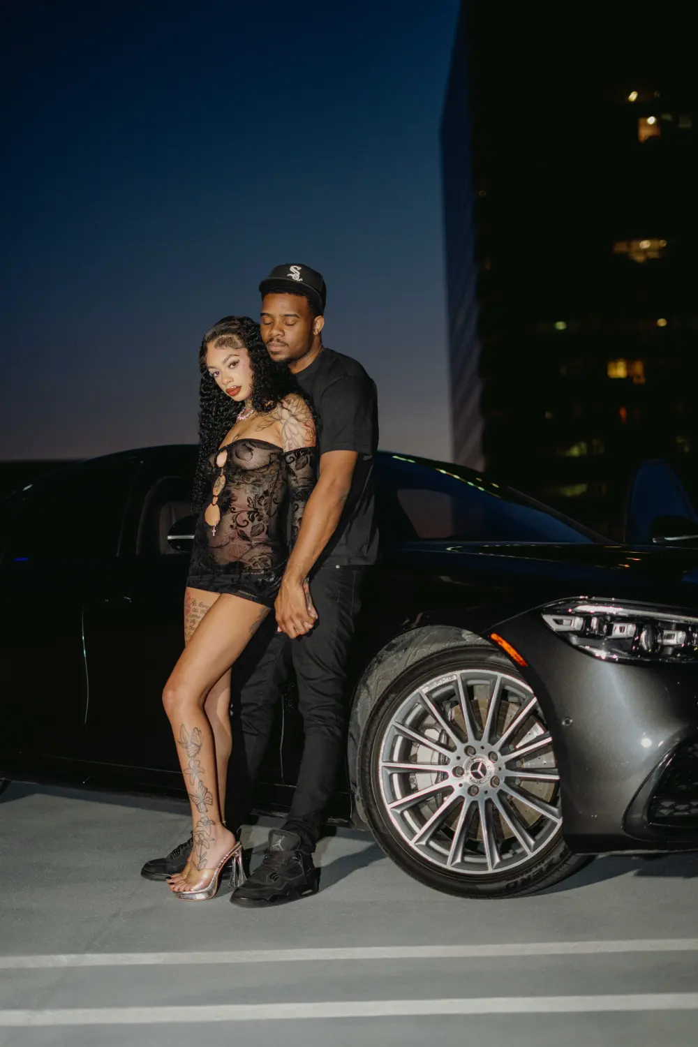 Couple embracing beside a black car on a rooftop at night, downtown towers behind them
