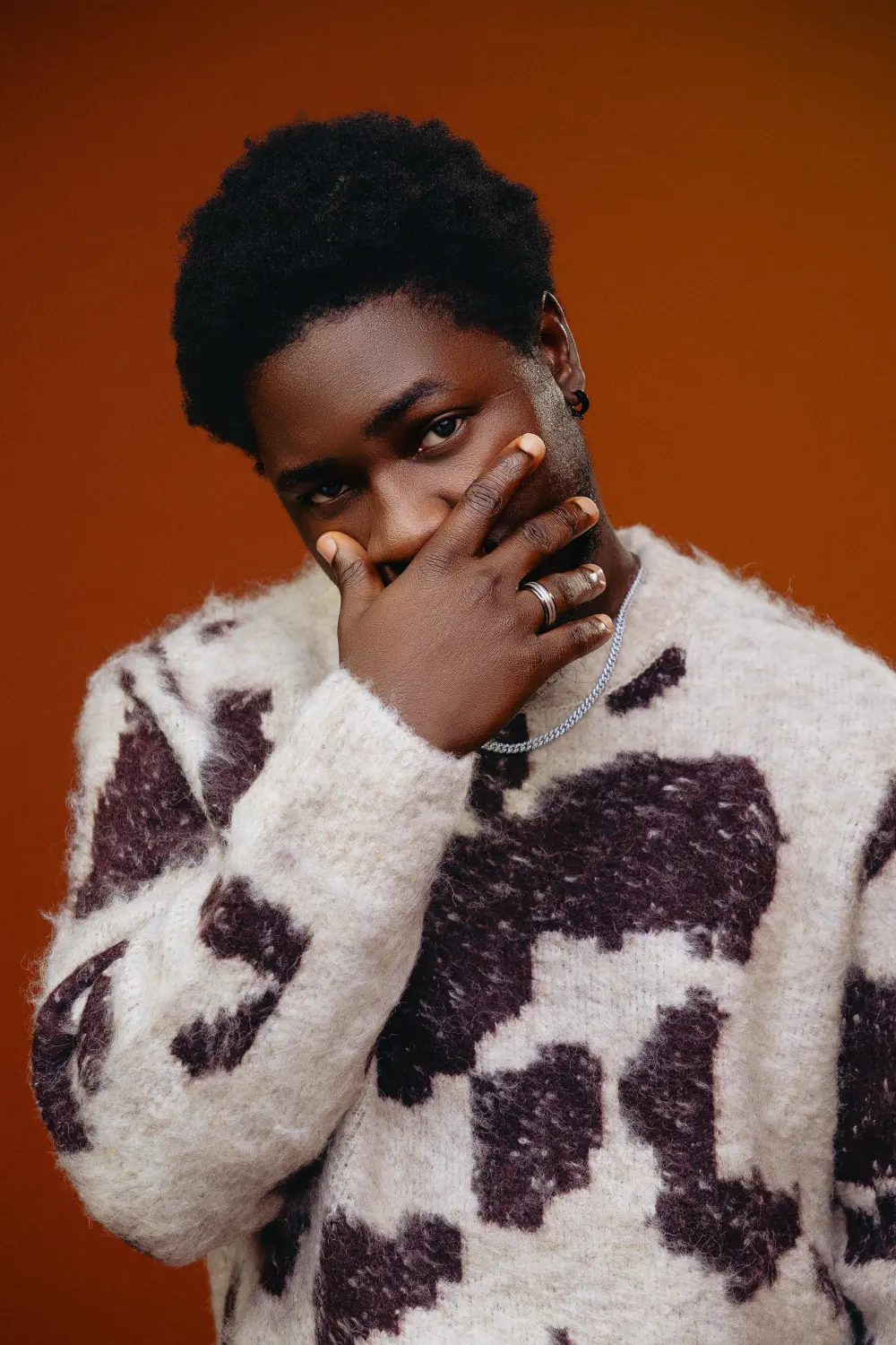 Man in a cow-print sweater with a hand over his mouth against a burnt orange backdrop