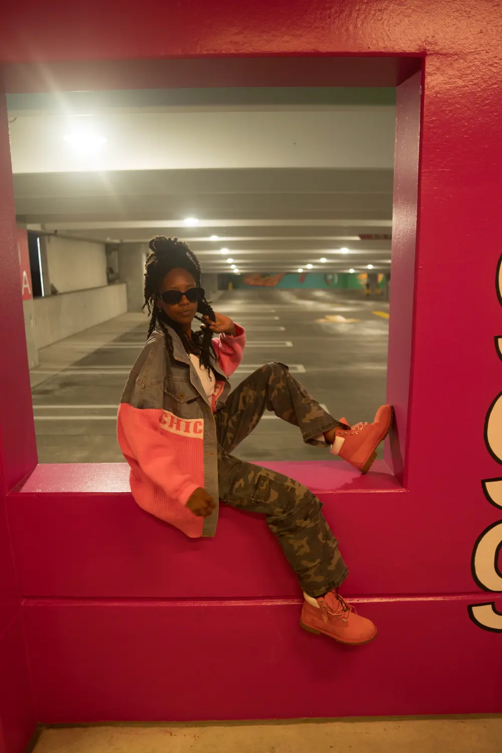 Woman in sunglasses, a pink jacket and camo pants sitting in a window cut into a pink parking garage wall
