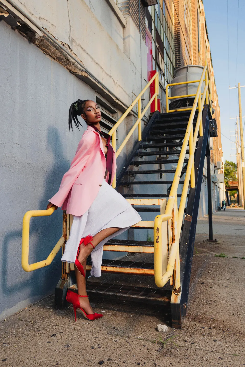 Woman in a pink blazer and red heels on a yellow-railed outdoor staircase in afternoon sun