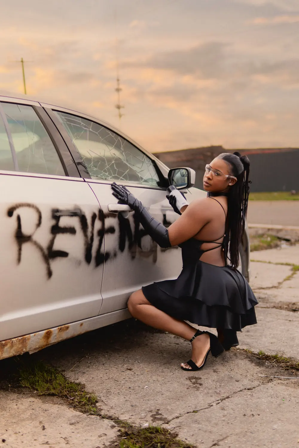 Woman in black gloves crouched with a spray can beside a car with REVENGE painted on the door