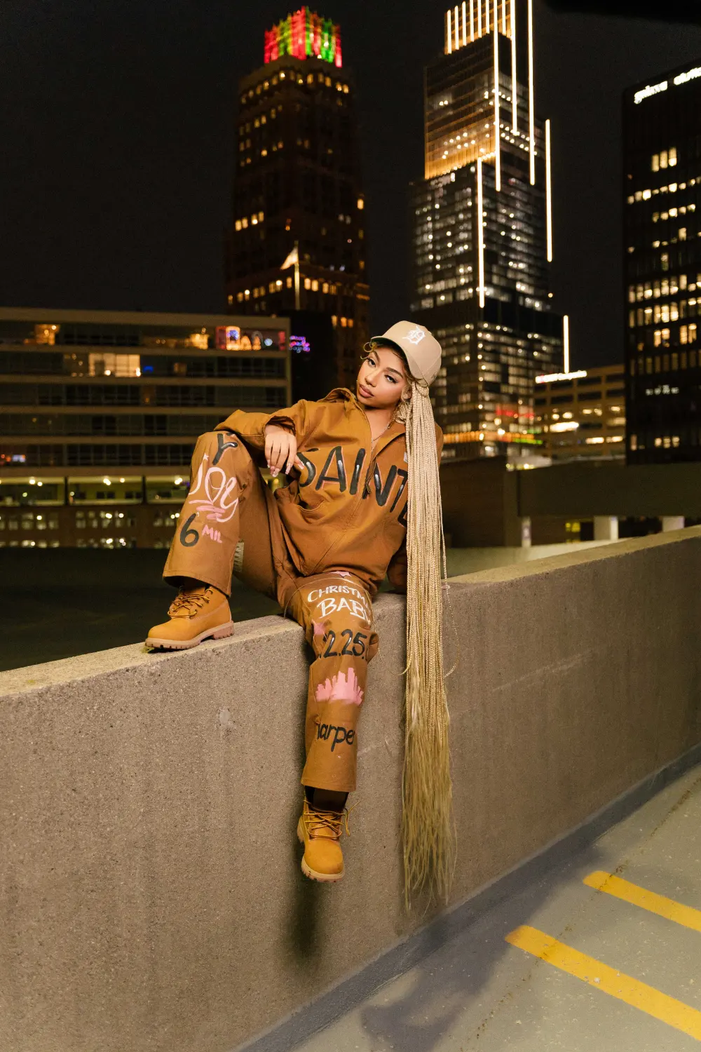 Woman in a brown outfit sitting on a rooftop ledge with lit-up downtown towers behind her at night