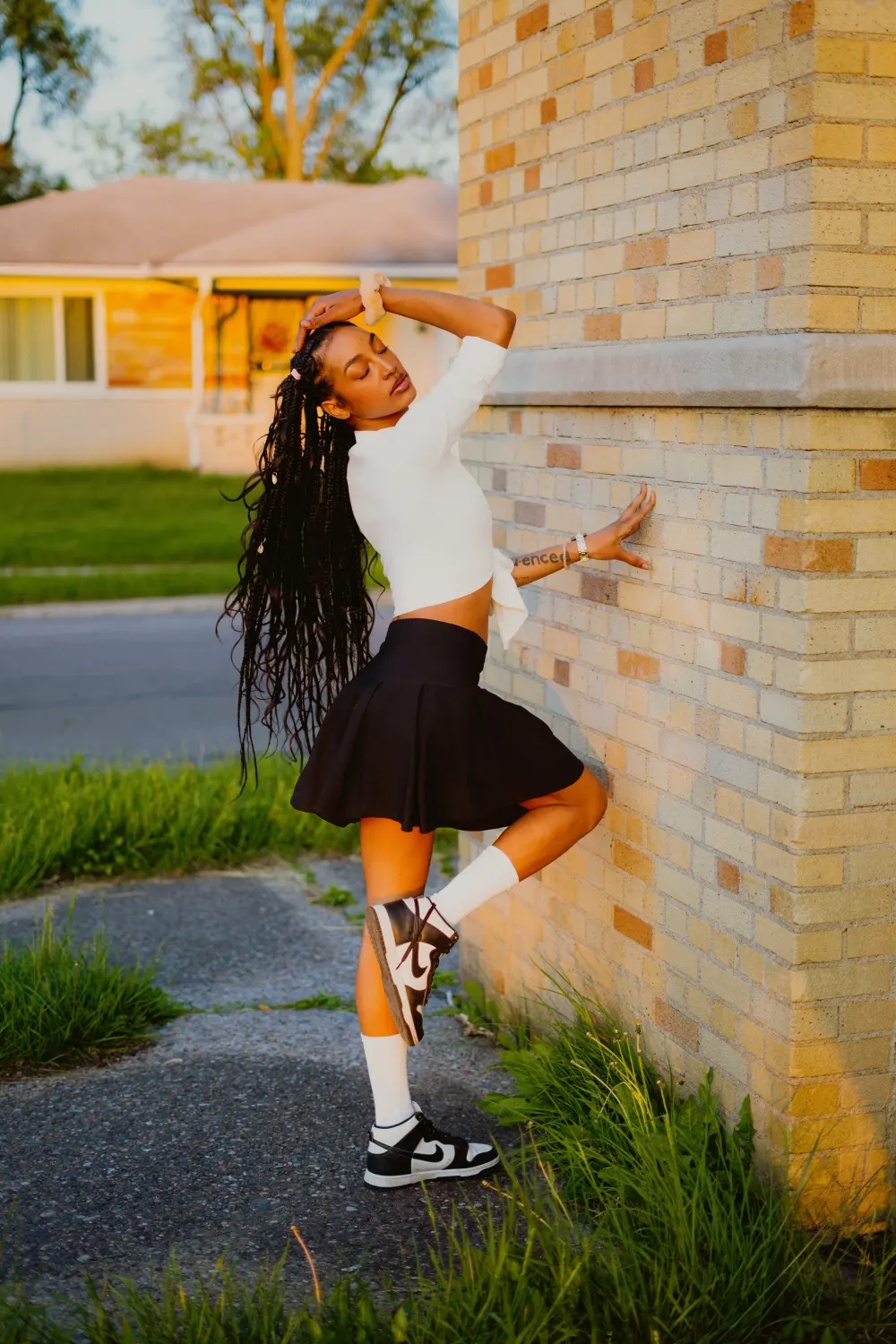 Woman in a white shirt and black skirt with one foot up against a brick wall in the evening sun
