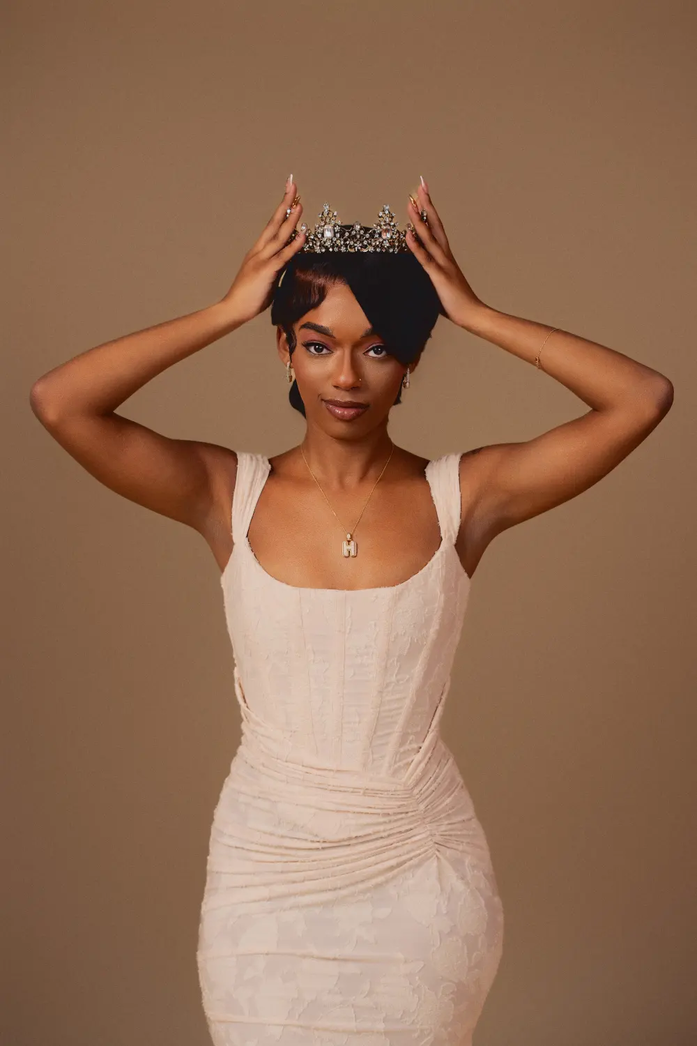 Woman in a white dress placing a crown on her head against a tan backdrop