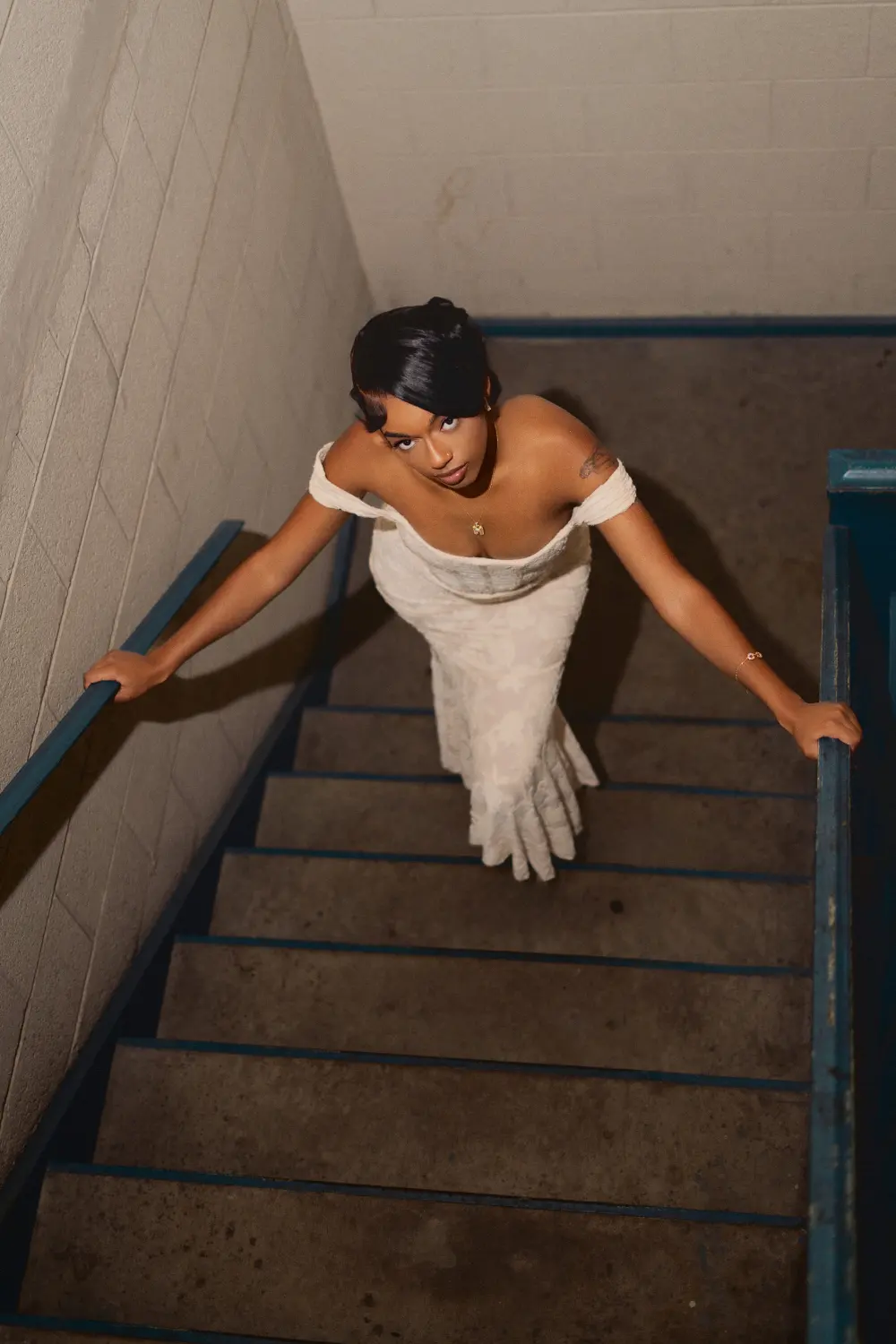 Woman in an off-shoulder cream gown leaning on the rails of a narrow stairwell, shot from above