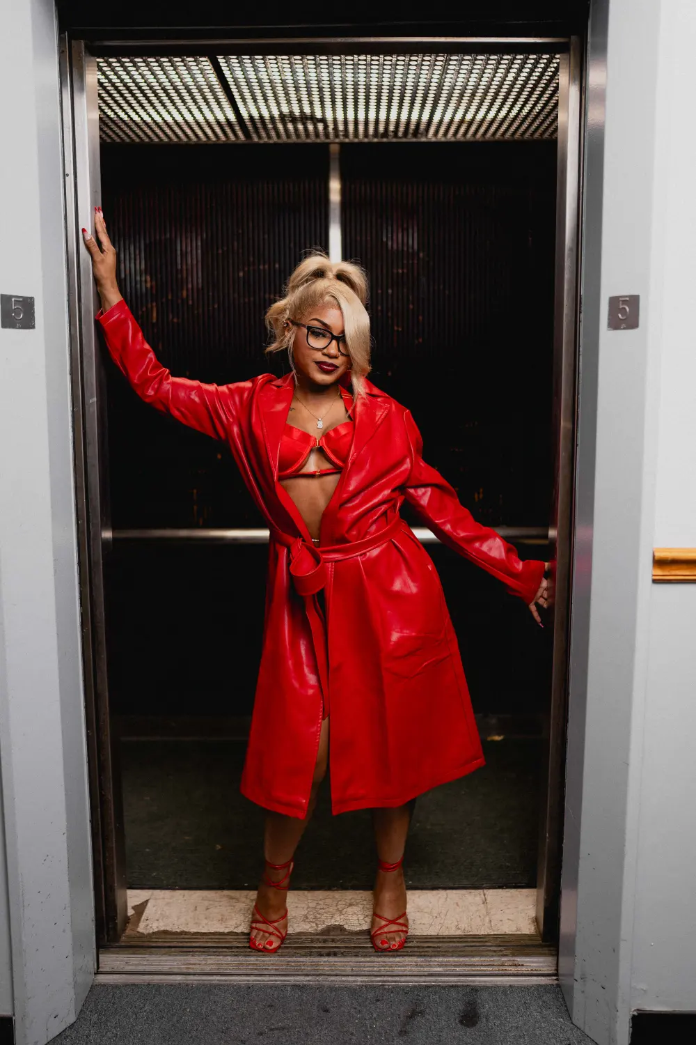 Woman in a red leather trench coat and glasses posing in an open freight elevator