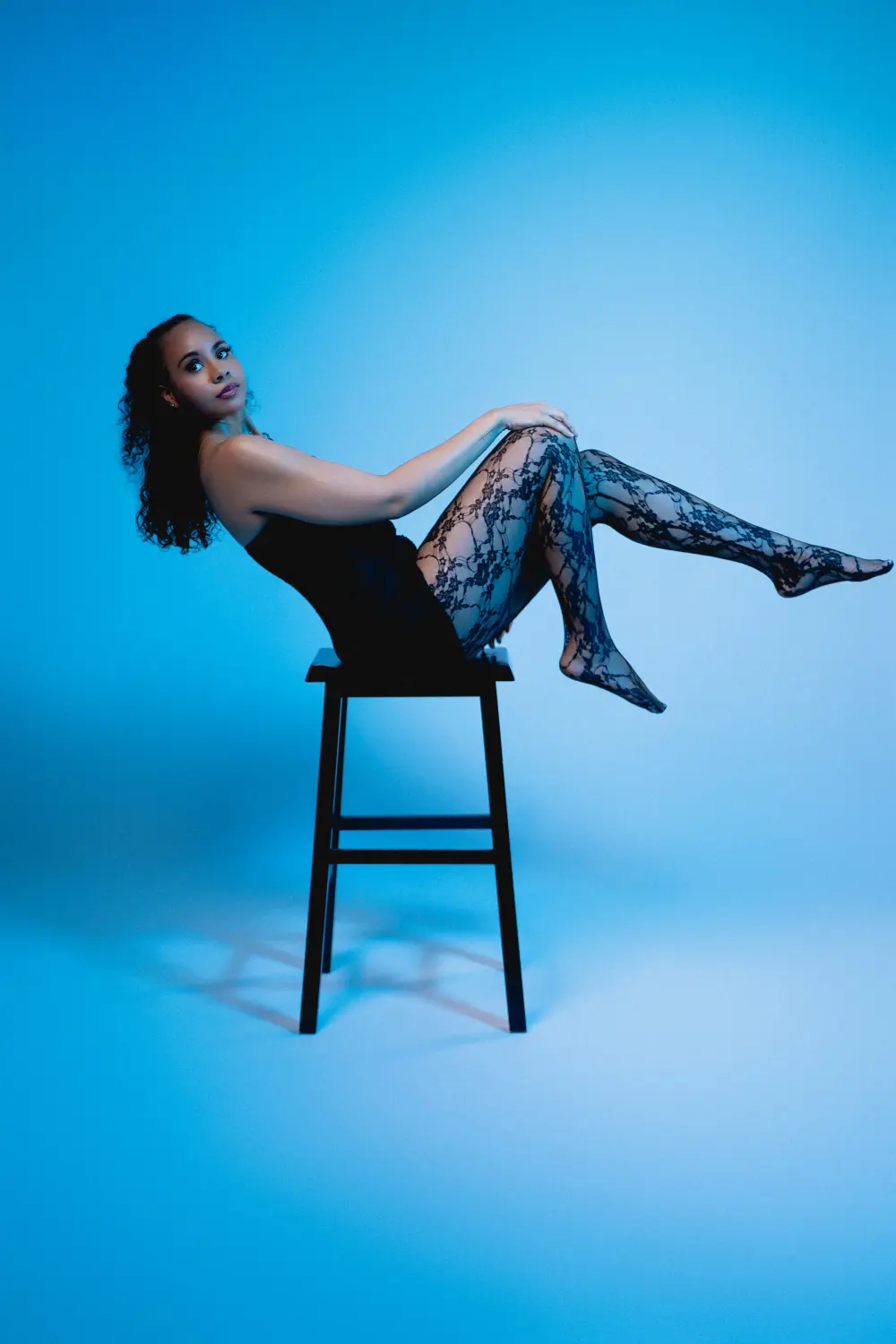 Woman in black lace tights sitting on a stool with her legs raised against a blue gel backdrop