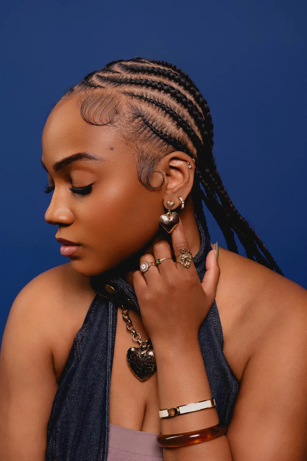 Close portrait of a woman with braided hair and gold jewelry, eyes down, against a deep blue backdrop