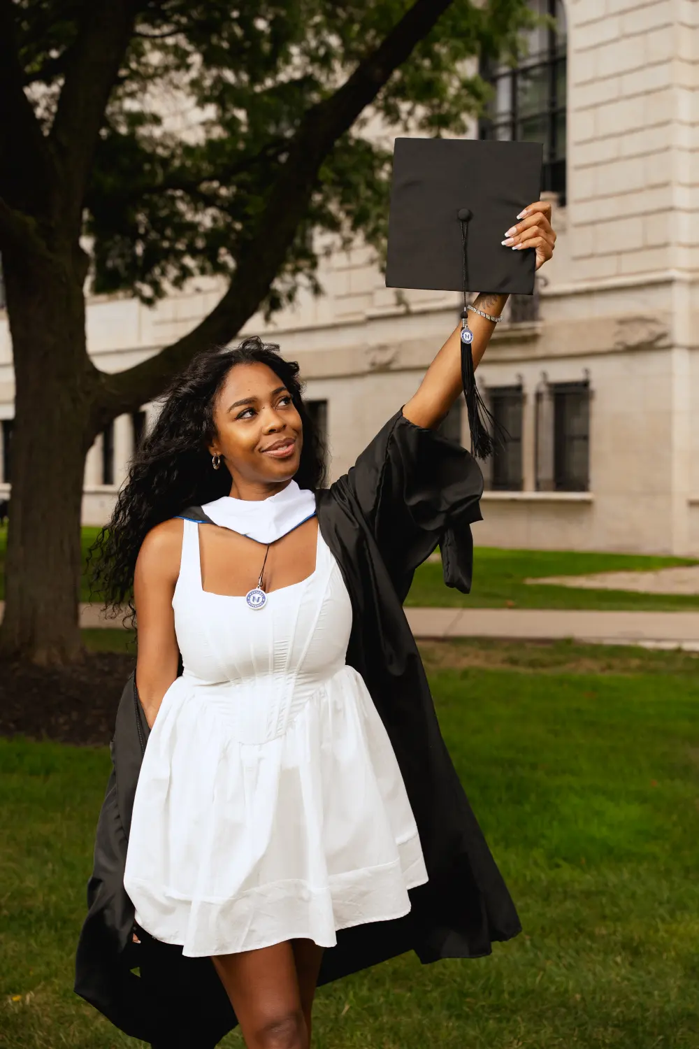 Graduate in a white dress and black gown holding her cap up under the trees