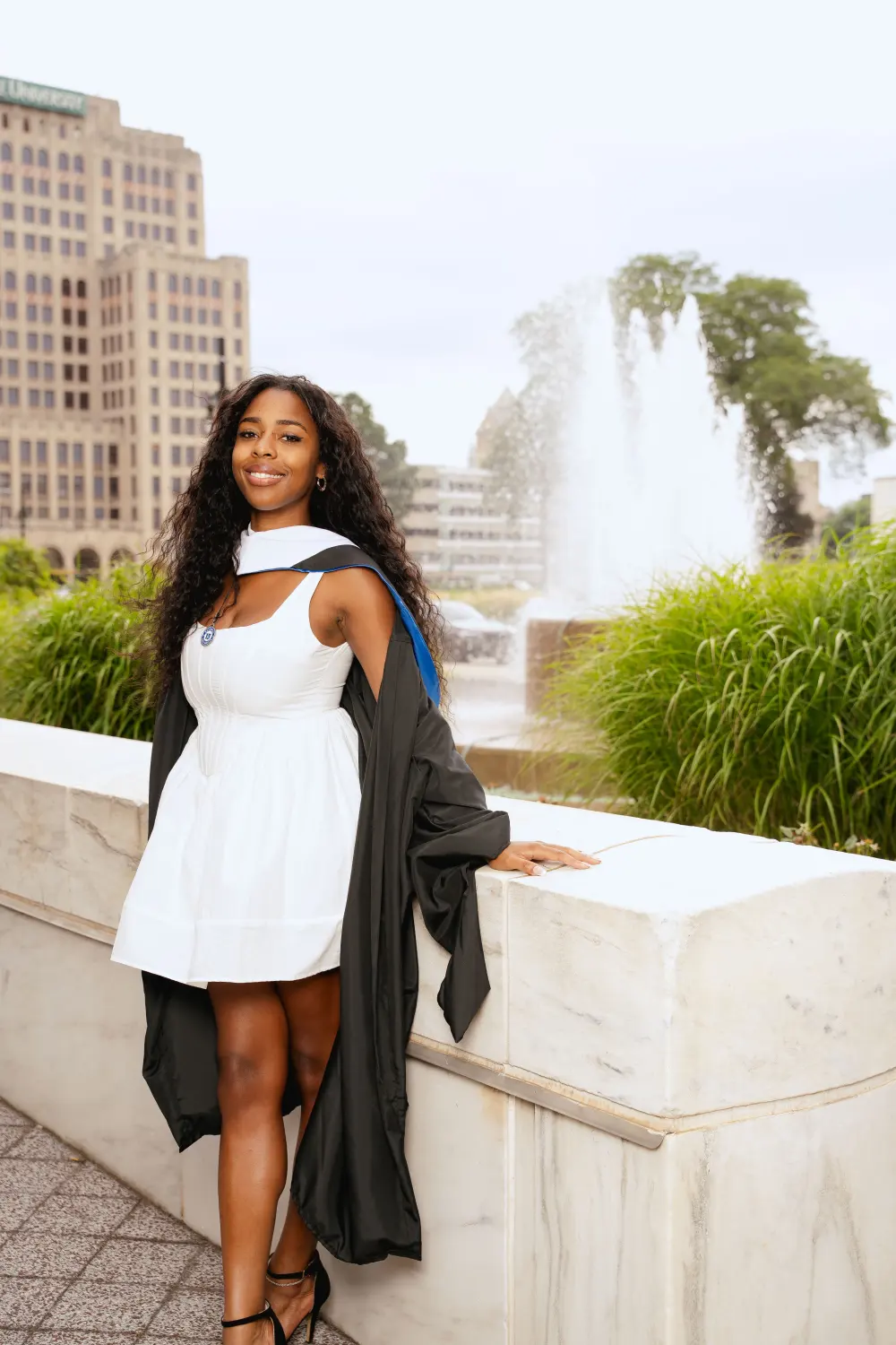 Graduate in a white dress with her gown off one shoulder beside a fountain, an office tower behind her