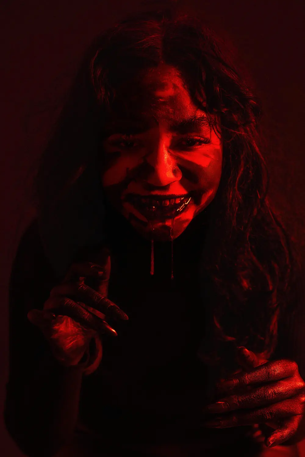 Woman lit red with a dark, smeared grin, reaching toward the camera with clawed fingers, her hair falling over her face