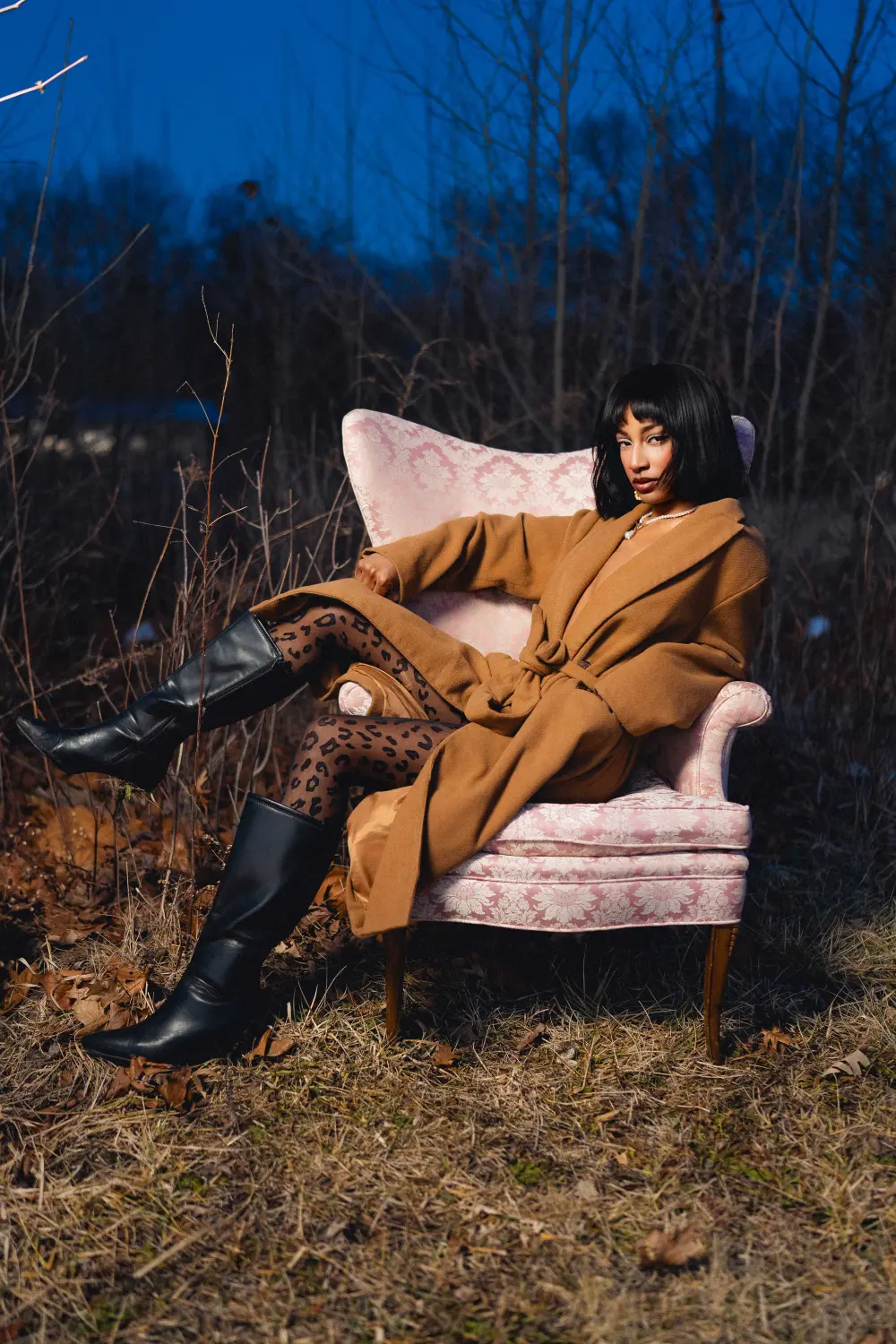Woman in a camel coat and black boots lounging sideways in a pink armchair among bare brush at dusk