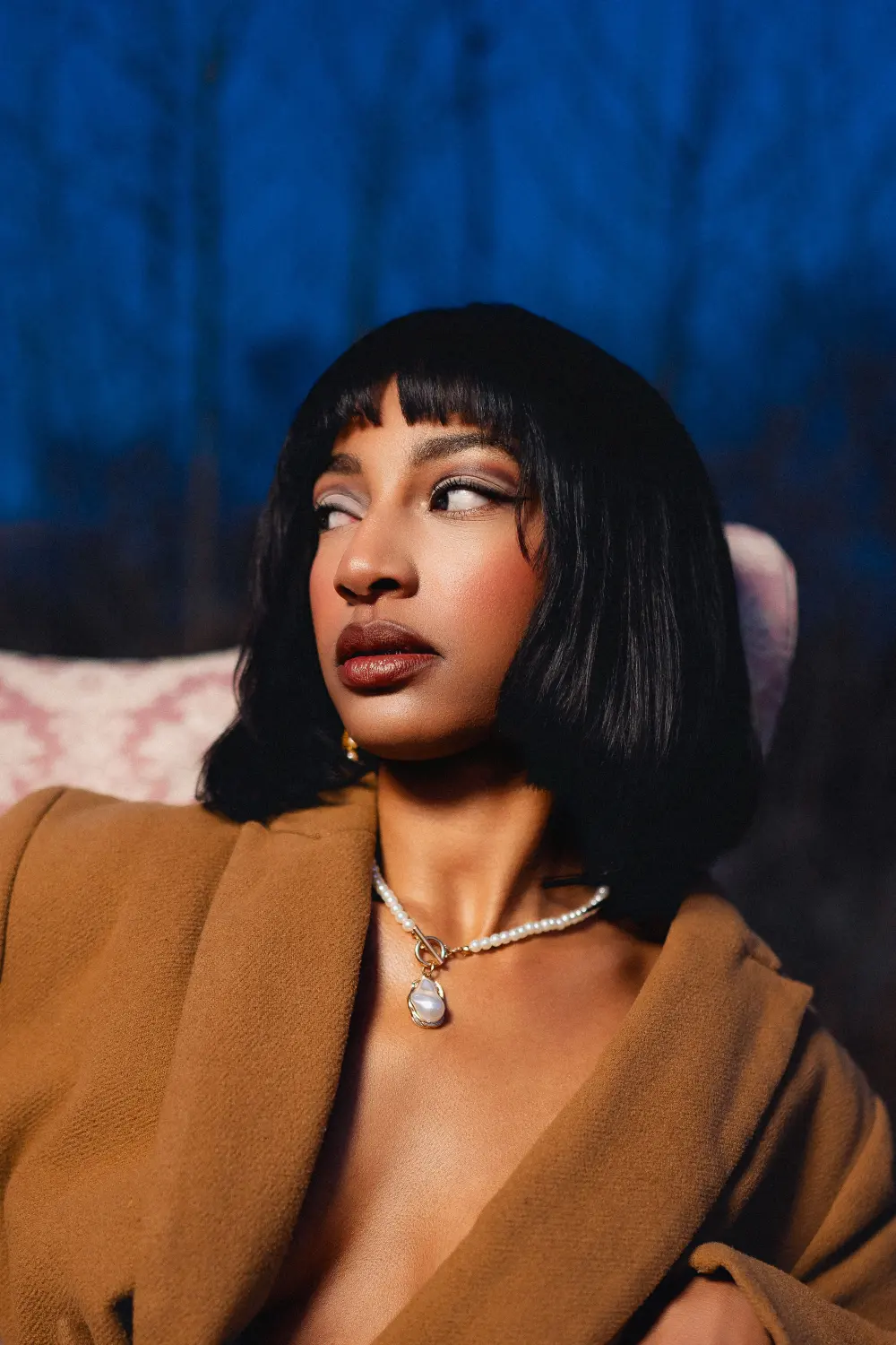 Close portrait of a woman with a black bob and a pearl necklace in a camel coat, the pink armchair behind her at blue hour