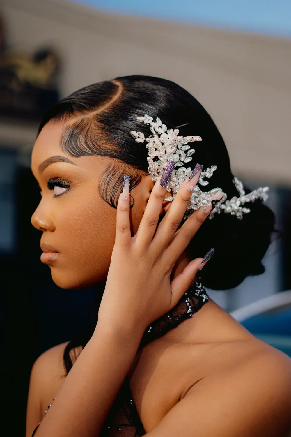 Close profile of a woman with a jeweled hairpiece and long nails, hand at her cheek