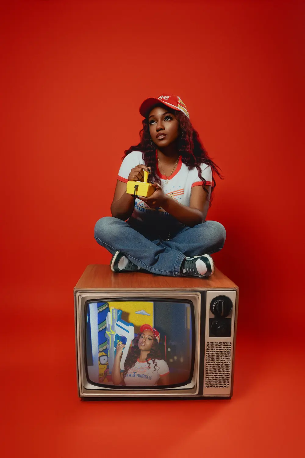 Woman in a red cap sitting cross-legged on top of an old TV against a red backdrop