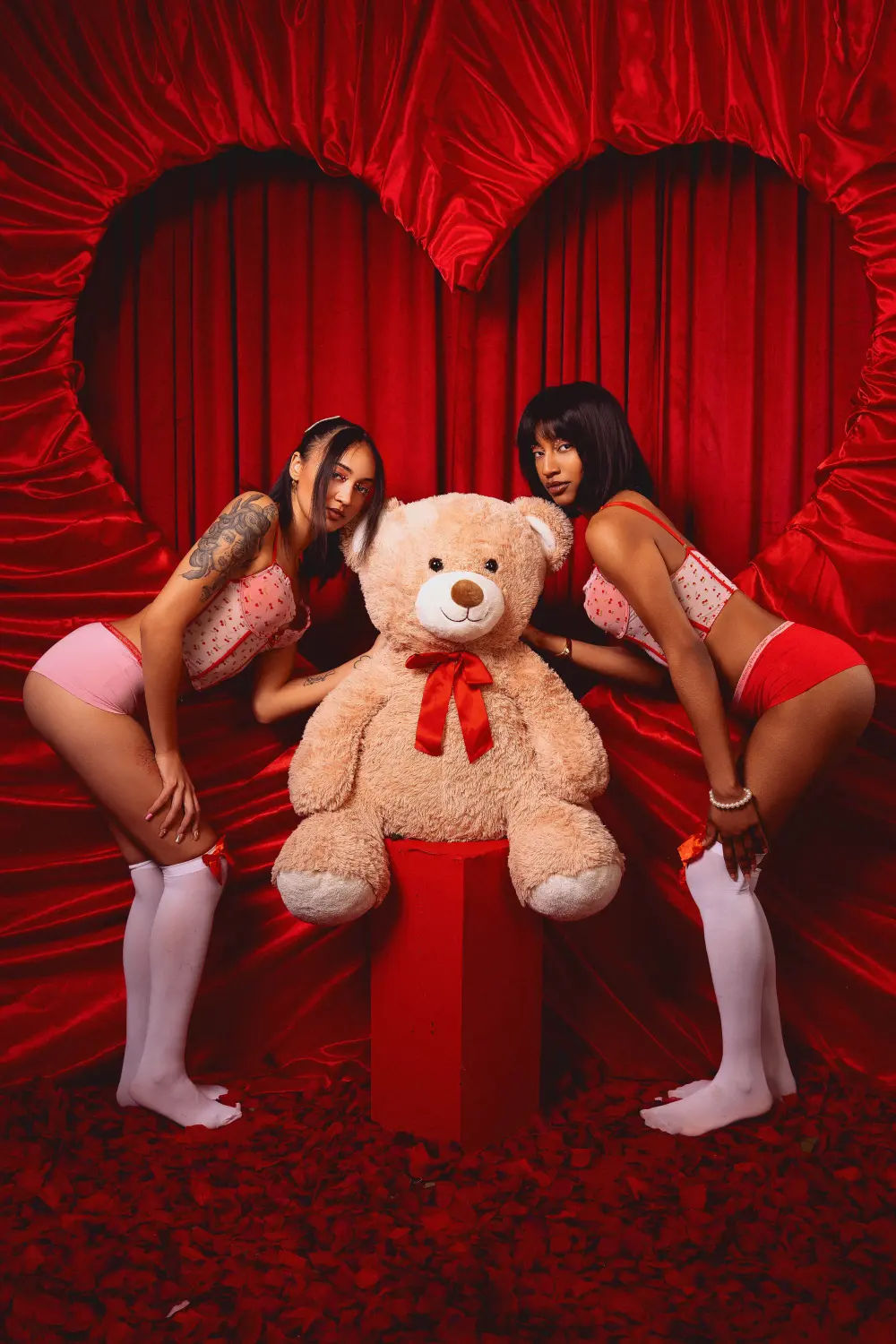 Two women in pink outfits and white stockings posing beside a giant teddy bear in a red heart set